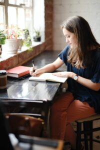 how do you write - woman sitting in front of black table writing on white book near window